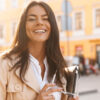 Cheerful brunette woman in jacket holding laptop computer and looking at the camera outdoors